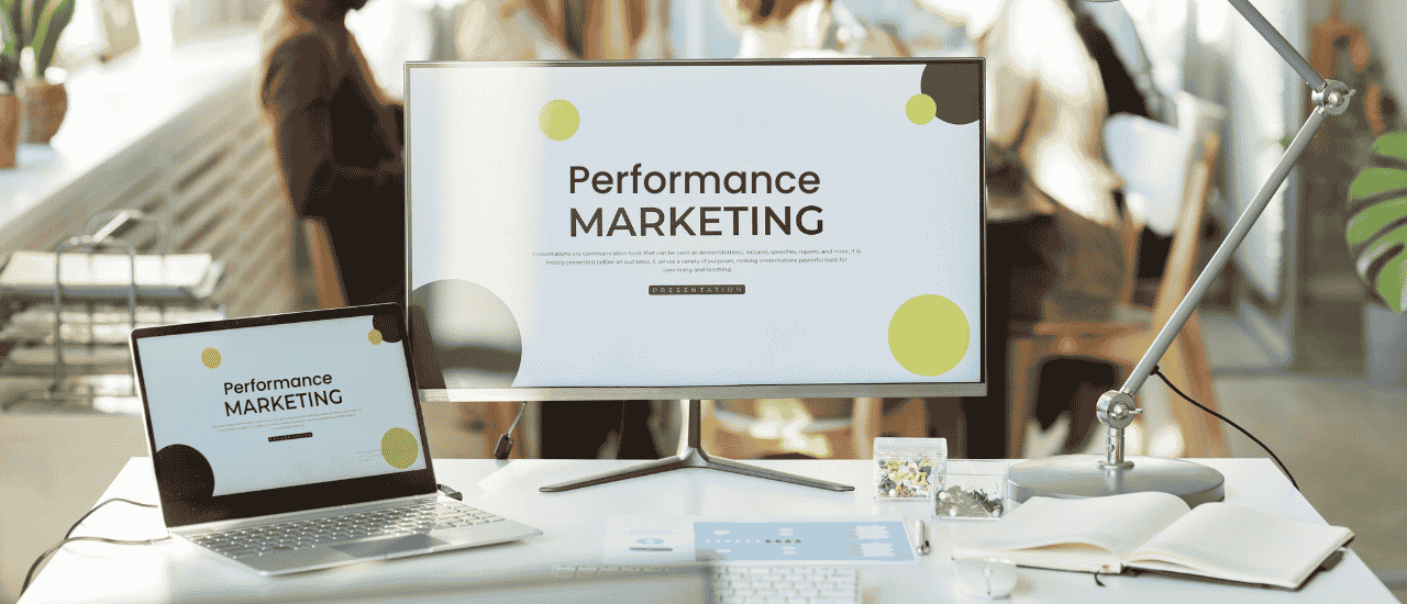 Moderner Arbeitsplatz mit Laptop und großem Monitor, auf beiden Bildschirmen ist die Präsentation „Performance Marketing“ zu sehen. Auf dem Schreibtisch befinden sich eine Lampe, ein geöffnetes Notizbuch, ein Stiftehalter und Büroutensilien. Im Hintergrund arbeiten unscharf dargestellte Personen in einem offenen Büro. Das Bild symbolisiert digitale Marketingstrategien, insbesondere im Bereich Performance Marketing und AutomateLeads.
